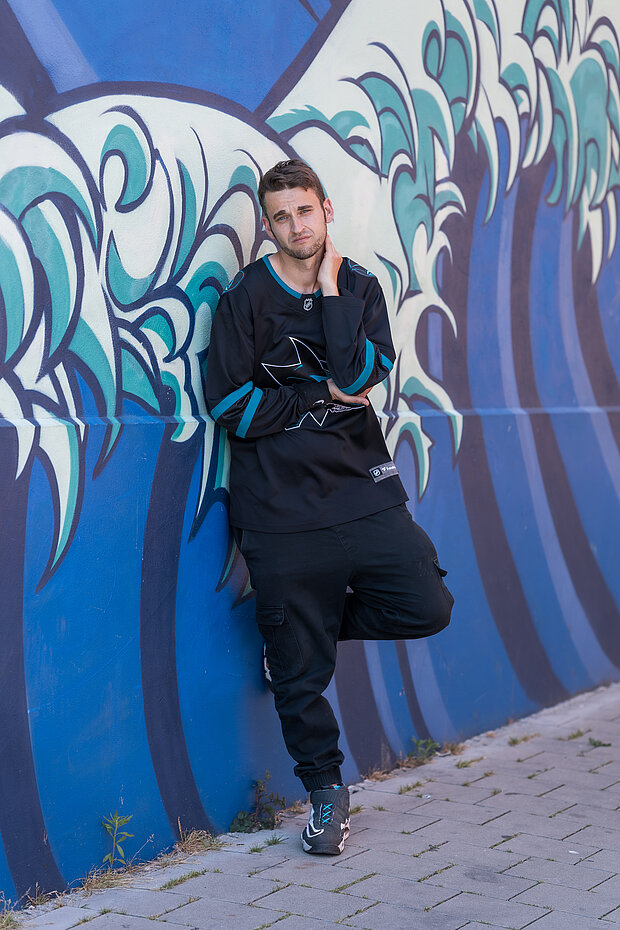 Ein junger Mann steht lässig an einer Wand, die mit blauen und grünen Wellenmuster bemalt ist. Er trägt ein schwarzes Sporttrikot und schwarze Hosen, und sieht nachdenklich aus. Der Hintergrund ist farbenfroh und hebt die Figur hervor.