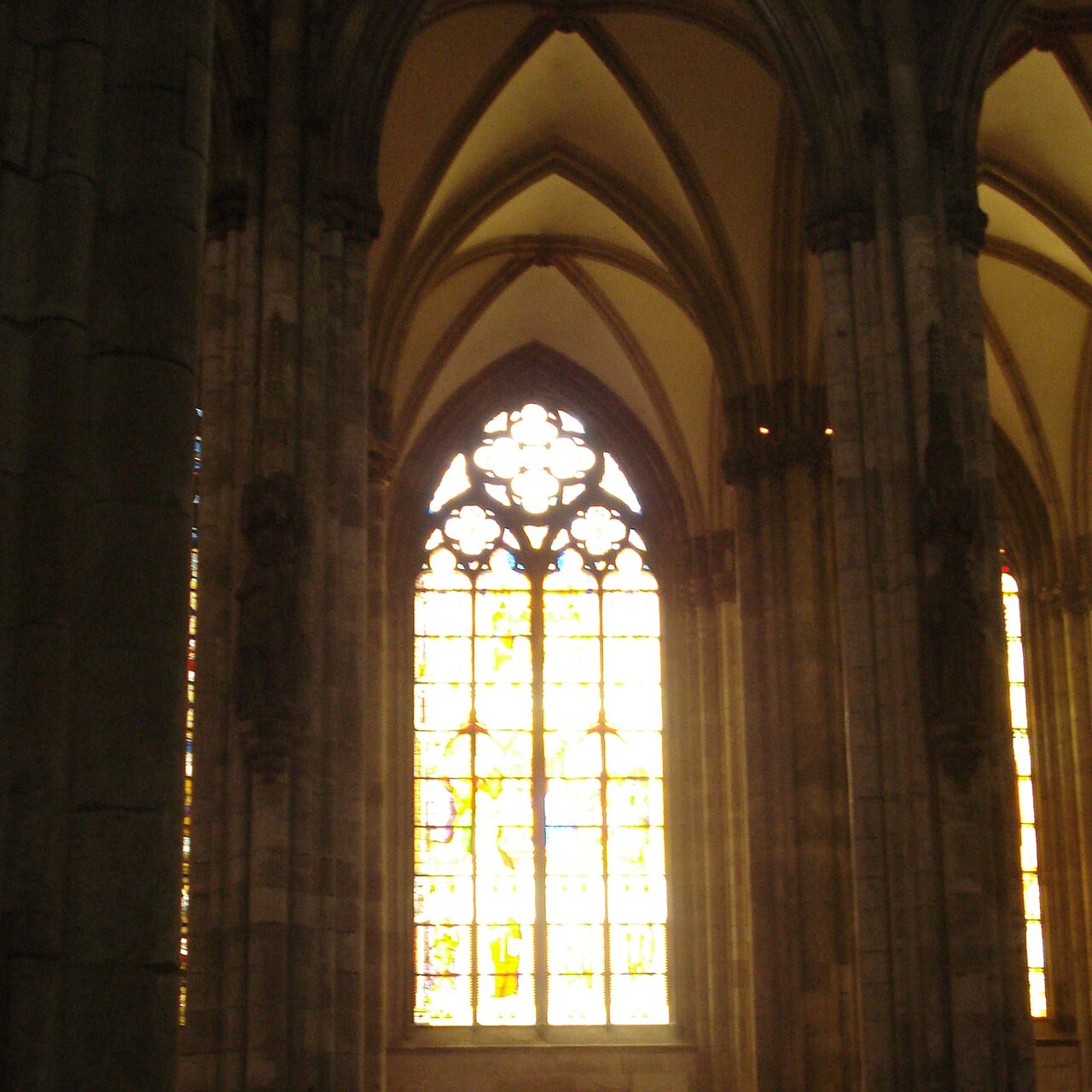 Ein lichtdurchflutetes Kirchenschiff mit hohen, gotischen Säulen. Rechts befindet sich ein großes, buntes Glasfenster, das Szenen oder Figuren zeigt, während die Wände und Säulen aus Stein die Höhe und Weite des Raumes betonen.